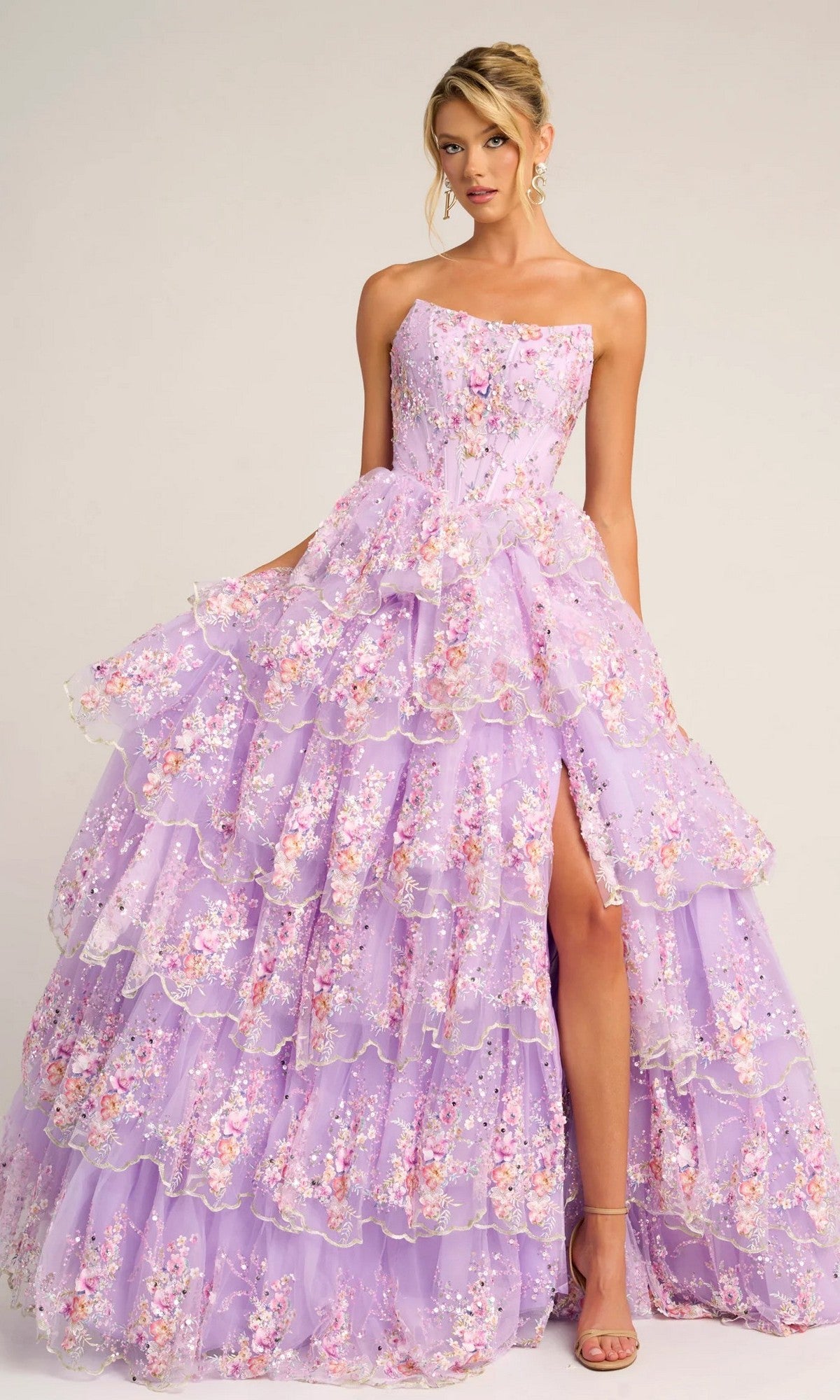 A woman wears the Portia and Scarlett PS26051 strapless floral ball gown in lilac, featuring tiered sheer layers, a high slit, and delicate embroidered floral appliqué.
